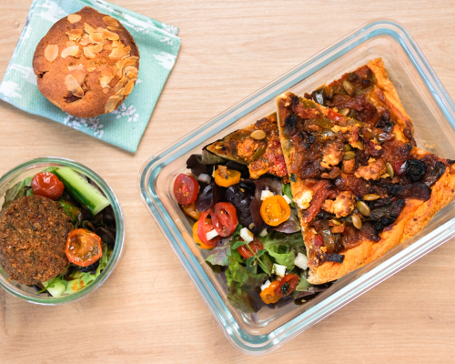 Un tupperware avec une quiche, des crudités, une entrée et un muffin aux amandes